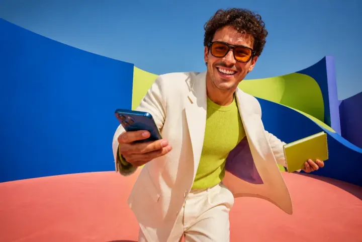 A founder in a cream suit moving toward the camera with a phone and notebook in a vivid blue, lime and coral studio